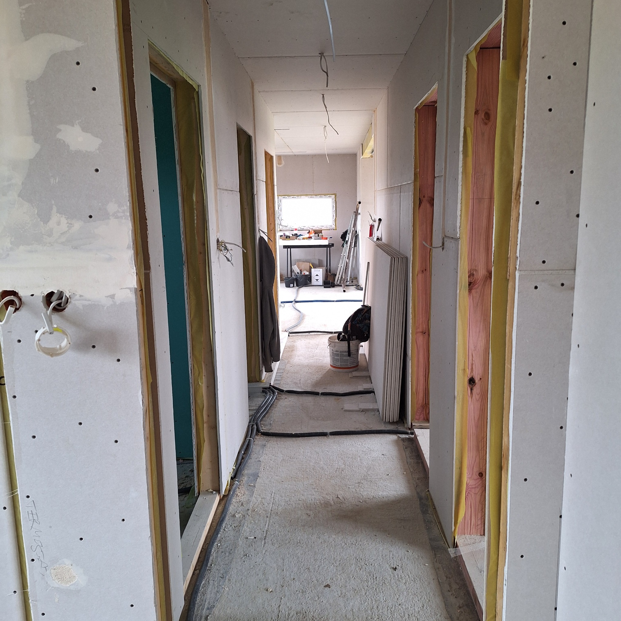 Korytarz w trakcie prac wykończeniowych: widoczne płyty karton-gips na ścianach, drewniane ramy drzwiowe z izolacją i instalacje elektryczne. W tle drabina i narzędzia.