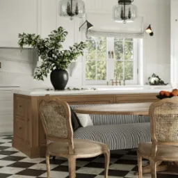 Elegancka, jasna kuchnia z czarno-białą podłogą w szachownicę, drewnianym stołem z siedziskiem w paski i bukietem w wazonie na blacie. Dwie krzesła z wiklinowymi oparciami.