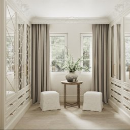 Mala & Co. Malwina Wilczek - Elegancka garderoba w jasnych barwach z lustrzanymi drzwiami, okna z zasłonami, stolik z kwiatami i pufami. Klasyczny, luksusowy styl.