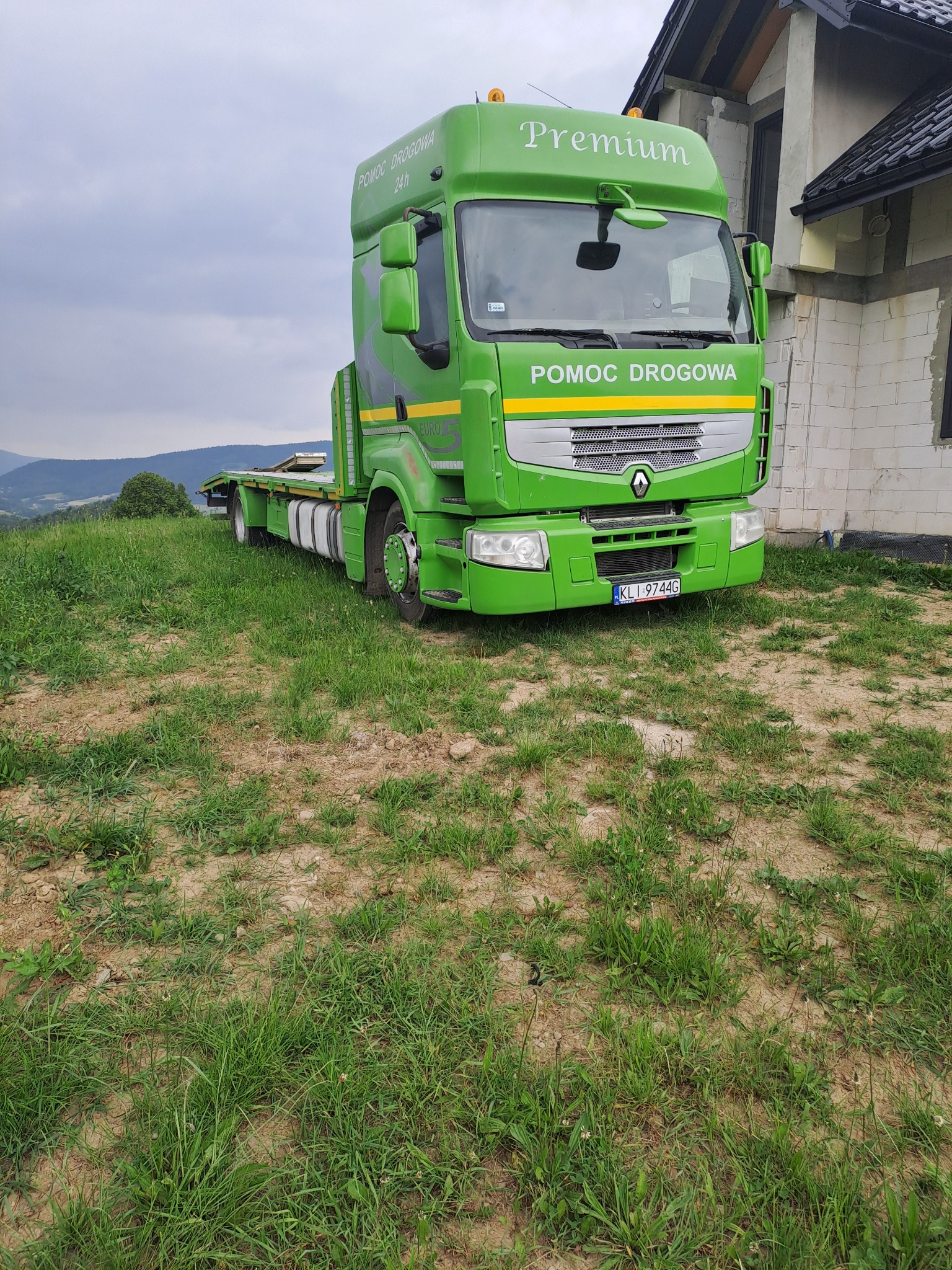 Zielona laweta Renault Premium z napisem 'Pomoc Drogowa' stoi na trawie, obok budynku w trakcie budowy. Widok na góry w tle, pochmurne niebo.