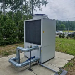 Duży, biały agregat wody lodowej z podłączonymi rurami na betonowej podstawie, otoczony zielenią i drzewami. Widoczne elementy instalacji i wentylator na górze.