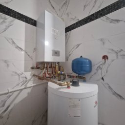 FJ Instal Jakub Furczyk - Biały piec gazowy z niebieskim zbiornikiem wyrównawczym na tle marmurowej ściany z czarnym pasem dekoracyjnym. Widoczne rury i zawory instalacji grzewczej.