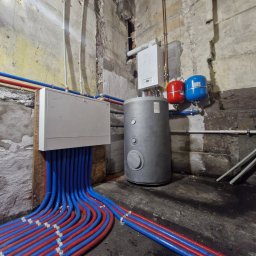 FJ Instal Jakub Furczyk - Instalacja CO w piwnicy: rozdzielacz, zbiornik, piec gazowy. Rury grzewcze prowadzone wzdłuż ściany. Widoczne elementy instalacji i surowy stan pomieszczenia.