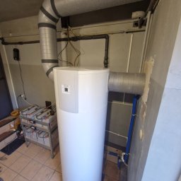 FJ Instal Jakub Furczyk - Biały, cylindryczny piec z szarymi rurami wentylacyjnymi w pomieszczeniu z beżowymi płytkami. Widoczne przewody i instalacje. Na podłodze akumulatory.