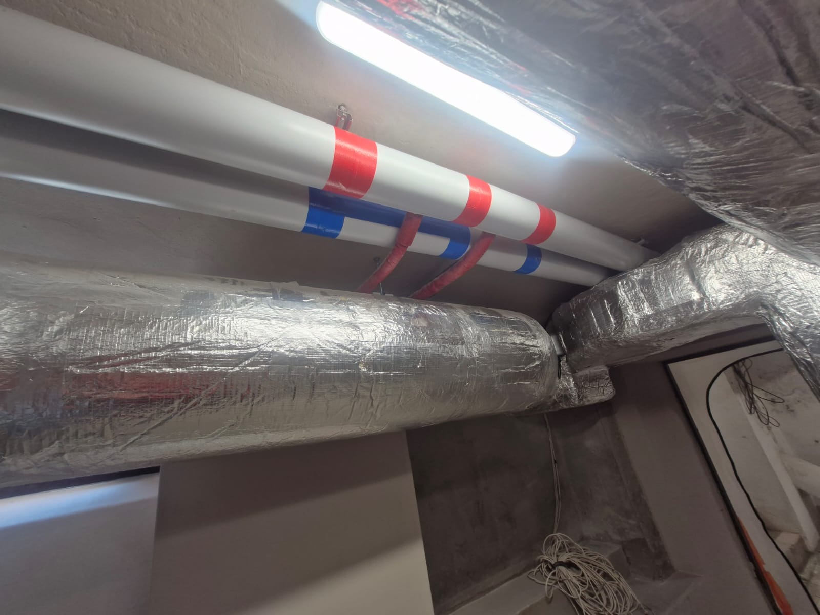 Izolowane rury wentylacyjne i grzewcze pod sufitem, z kolorowymi oznaczeniami, w pomieszczeniu technicznym. Widoczne kable i oświetlenie.