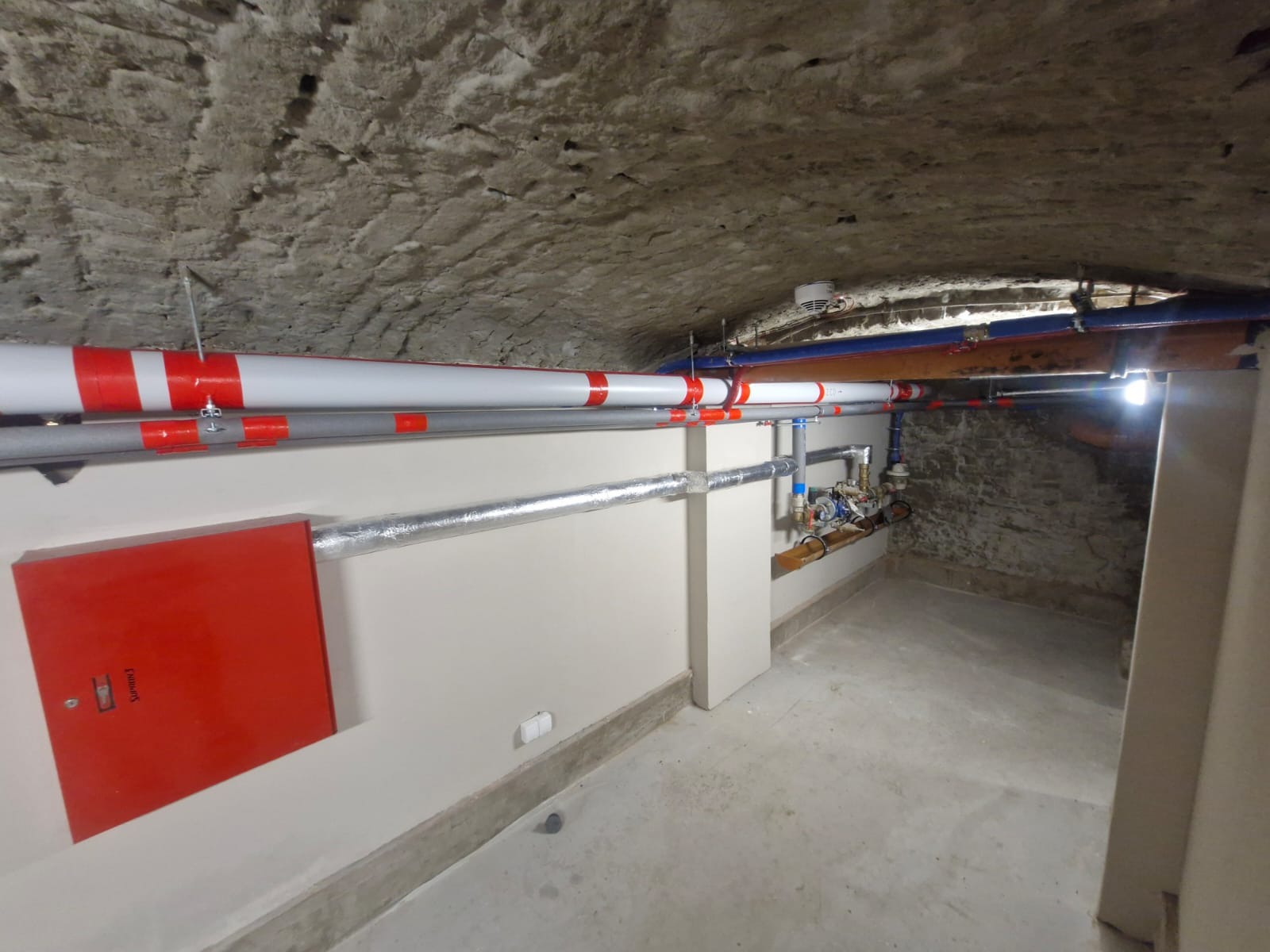 Instalacja rur i zaworów w piwnicy z kamiennym sufitem. Czerwona szafka na ścianie. Rury pomalowane w czerwono-białe pasy. Widoczne przyłącza i izolacja termiczna.
