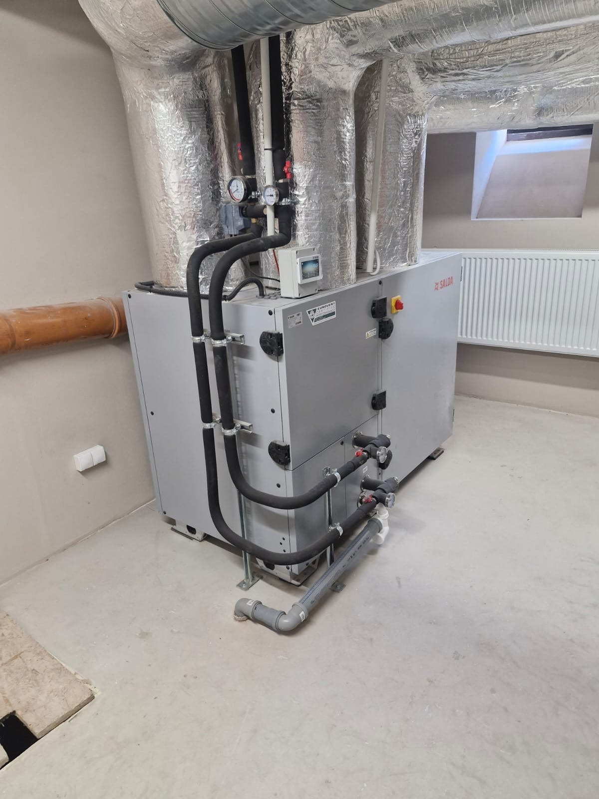 Szara centrala wentylacyjna Salda z czarnymi rurami w pomieszczeniu technicznym. Widoczne srebrne rury wentylacyjne pod sufitem i grzejnik żeberkowy w tle.