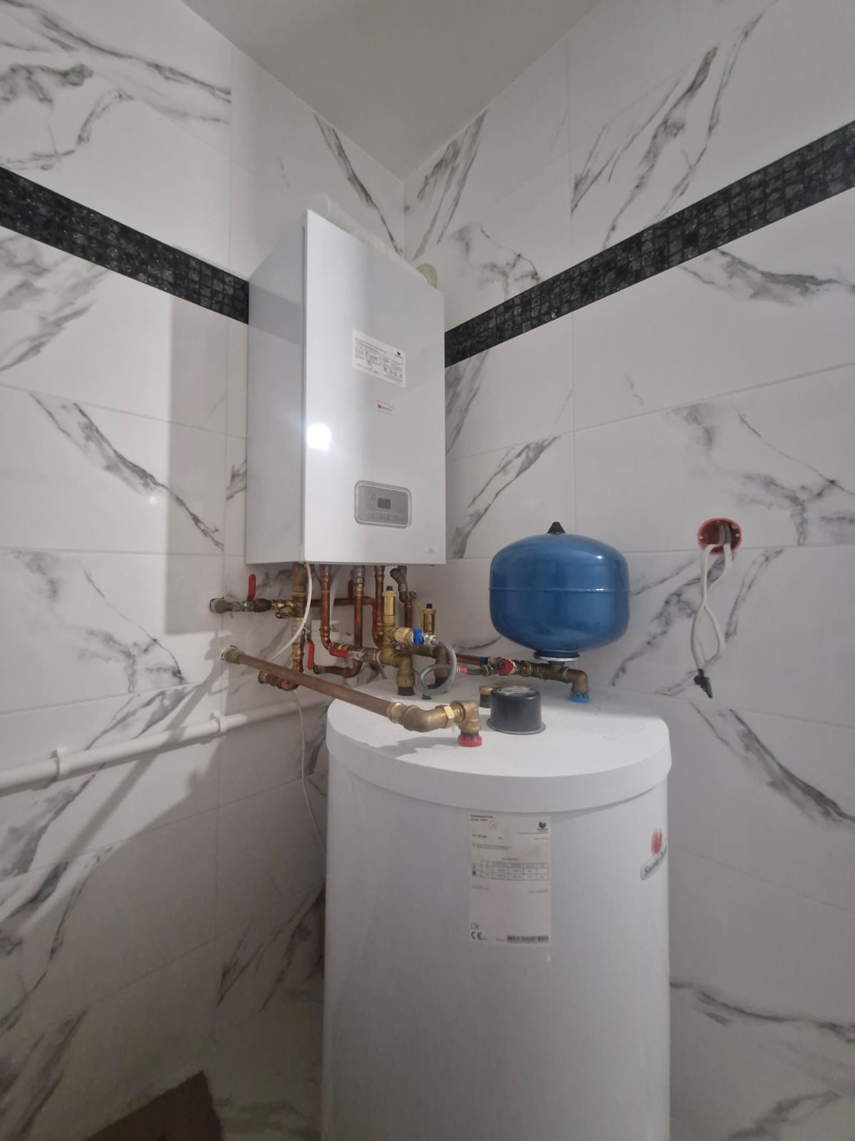 Biały piec gazowy z niebieskim zbiornikiem wyrównawczym na tle marmurowej ściany z czarnym pasem dekoracyjnym. Widoczne rury i zawory instalacji grzewczej.