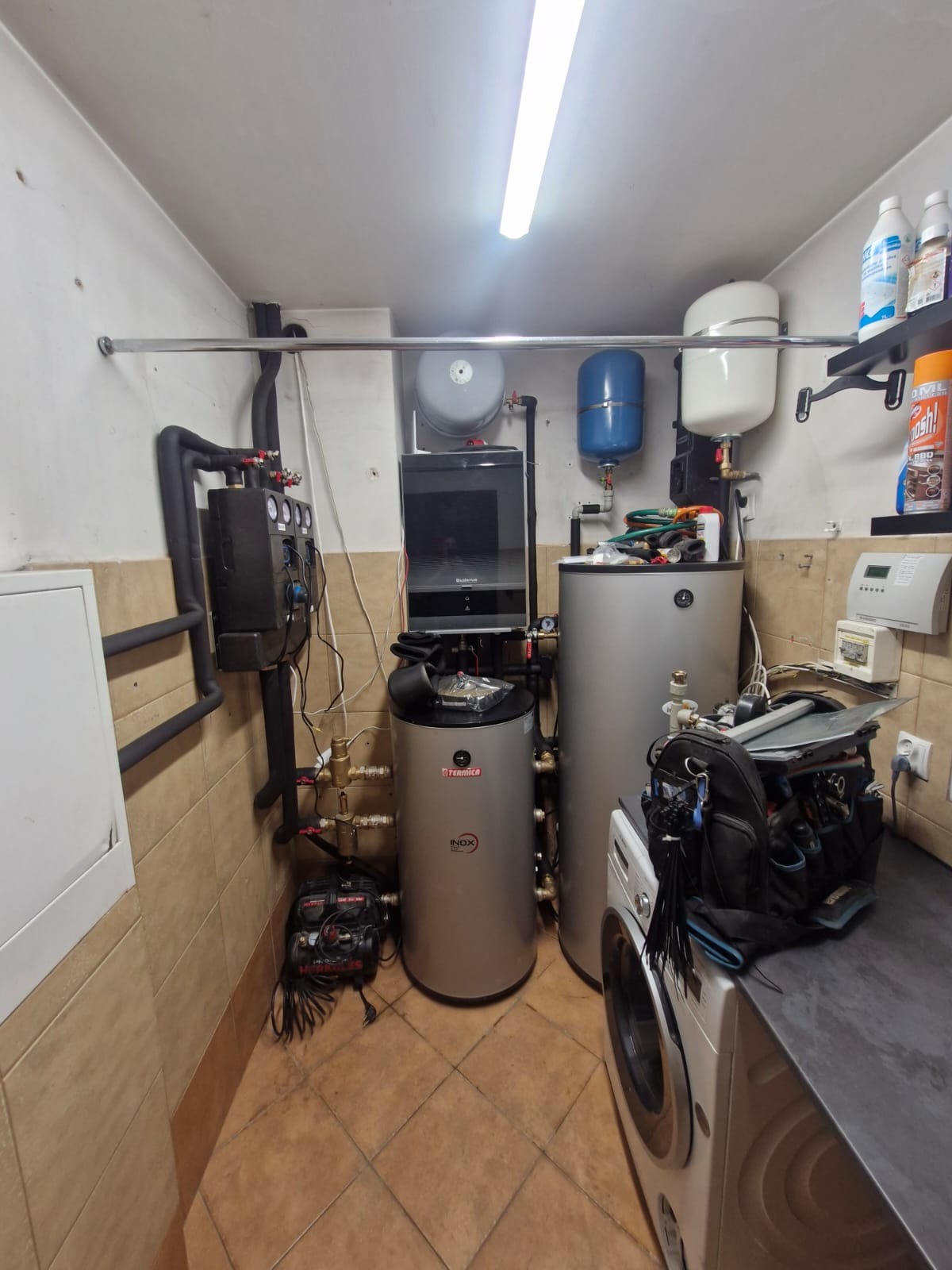 Dwa srebrne bojlery Termica Inox w kotłowni, obok pralka, narzędzia i instalacja CO. Widoczne rury, zawory i czujniki. Oświetlenie jarzeniowe.
