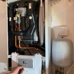 3CE Karol Lange - Ustawianie parametrów pieca gazowego wiszącego na ścianie. Widoczne rury miedziane, czarne elementy instalacji i zbiornik wyrównawczy. Ręka reguluje temperaturę.