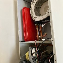 3CE Karol Lange - Wnętrze kotła gazowego z widocznymi elementami instalacji, zbiornikiem wyrównawczym i podzespołami. Widok z bliska na komponenty wewnątrz obudowy.