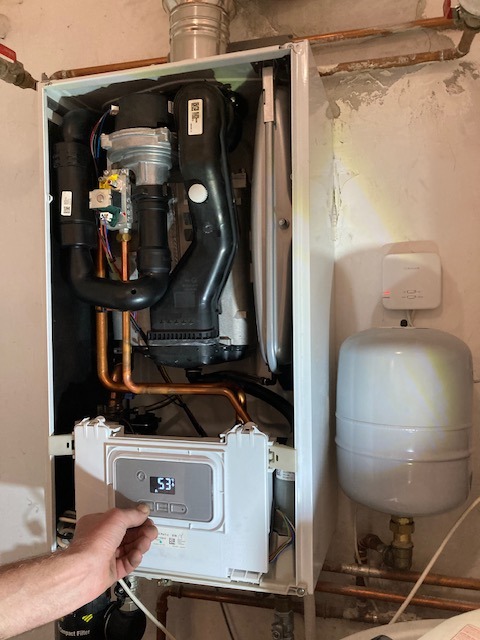 Ustawianie parametrów pieca gazowego wiszącego na ścianie. Widoczne rury miedziane, czarne elementy instalacji i zbiornik wyrównawczy. Ręka reguluje temperaturę.