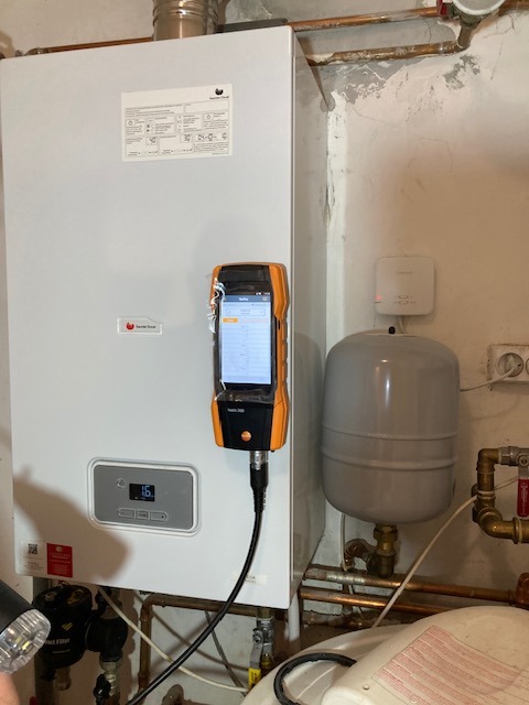 Biały piec gazowy z podłączonym analizatorem spalin Testo 300, widoczny zbiornik wyrównawczy i instalacja. Ściana z widocznymi śladami wilgoci.