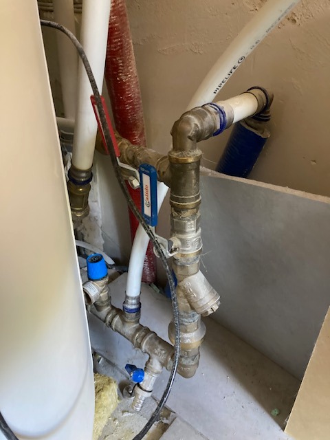 Fragment instalacji wodnej z zaworami i filtrem, widoczne rury z tworzywa i miedziane złączki. Tło to surowa ściana i fragment bojlera.