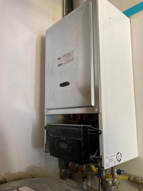 Biały, wiszący piec gazowy z czarnym panelem sterowania, widoczne rury i przyłącza. Piec zamontowany na jasnej, lekko zabrudzonej ścianie.