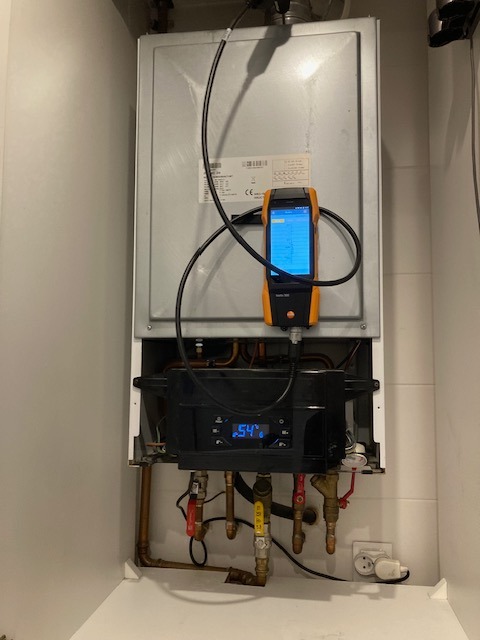 Piec gazowy w zabudowie, podłączony do analizatora spalin Testo 300, z widocznymi rurami i zaworami, wskazujący na etap diagnostyki lub serwisu.