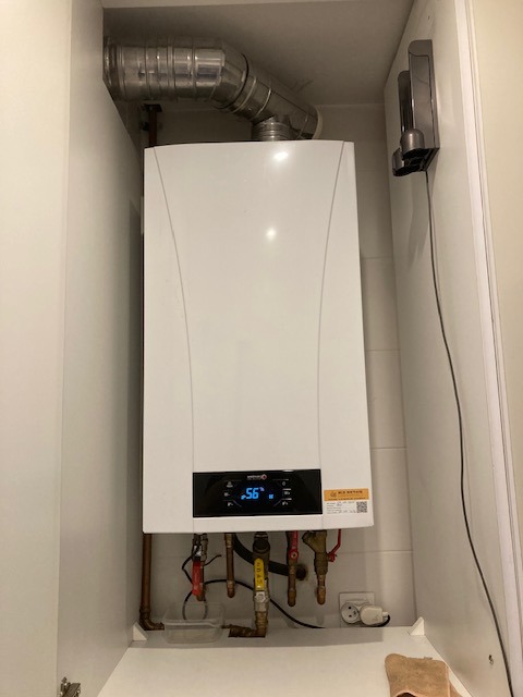 Biały piec gazowy zamontowany w szafce, widoczne rury doprowadzające, przewód kominowy i panel sterowania z wyświetlaną temperaturą.