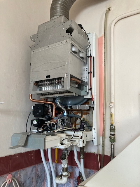 Otwarty piec gazowy w trakcie serwisu. Widoczne elementy wewnętrzne, miedziane rurki i przewody. Tło stanowią ściany z fragmentami płytek.