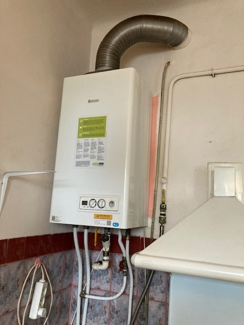 Biały piec gazowy Beretta z podłączonymi rurami, zamontowany na ścianie w pomieszczeniu z okapem i płytkami. Widoczny przewód kominowy.