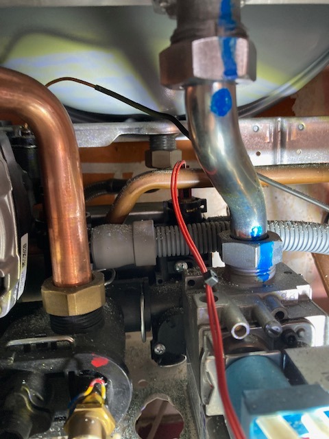 Wnętrze kotła gazowego z miedzianymi rurami i zaworami, widoczne przewody elektryczne i elementy instalacji. Detal z niebieskimi oznaczeniami na metalowych złączkach.