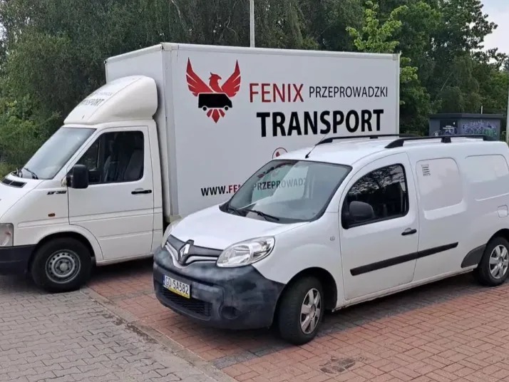 Dwa białe samochody dostawcze firmy Fenix Przeprowadzki Transport, zaparkowane na kostce brukowej, z zielonym tłem drzew.