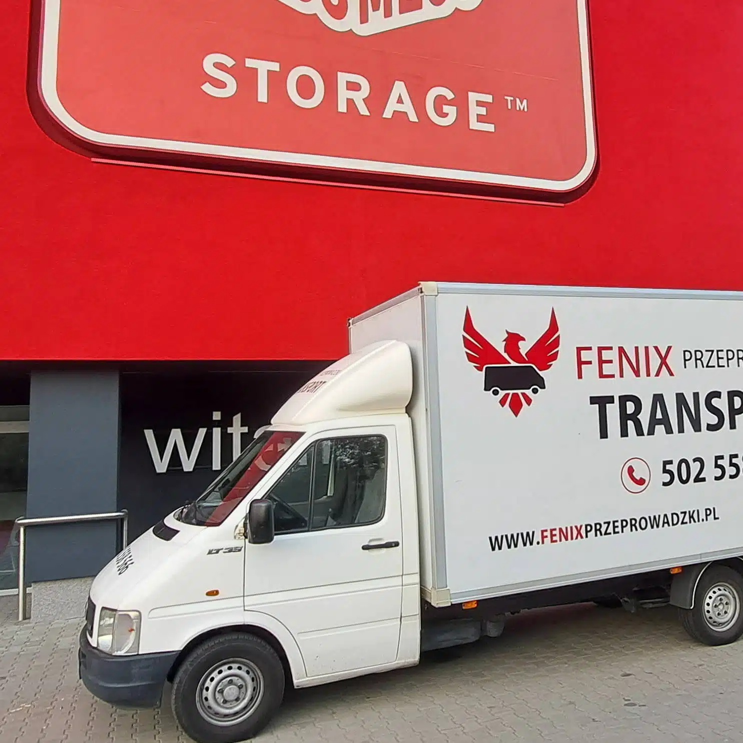 Biała furgonetka z logo Fenix Przeprowadzki na tle czerwonego budynku magazynowego, prezentująca usługi transportowe firmy.