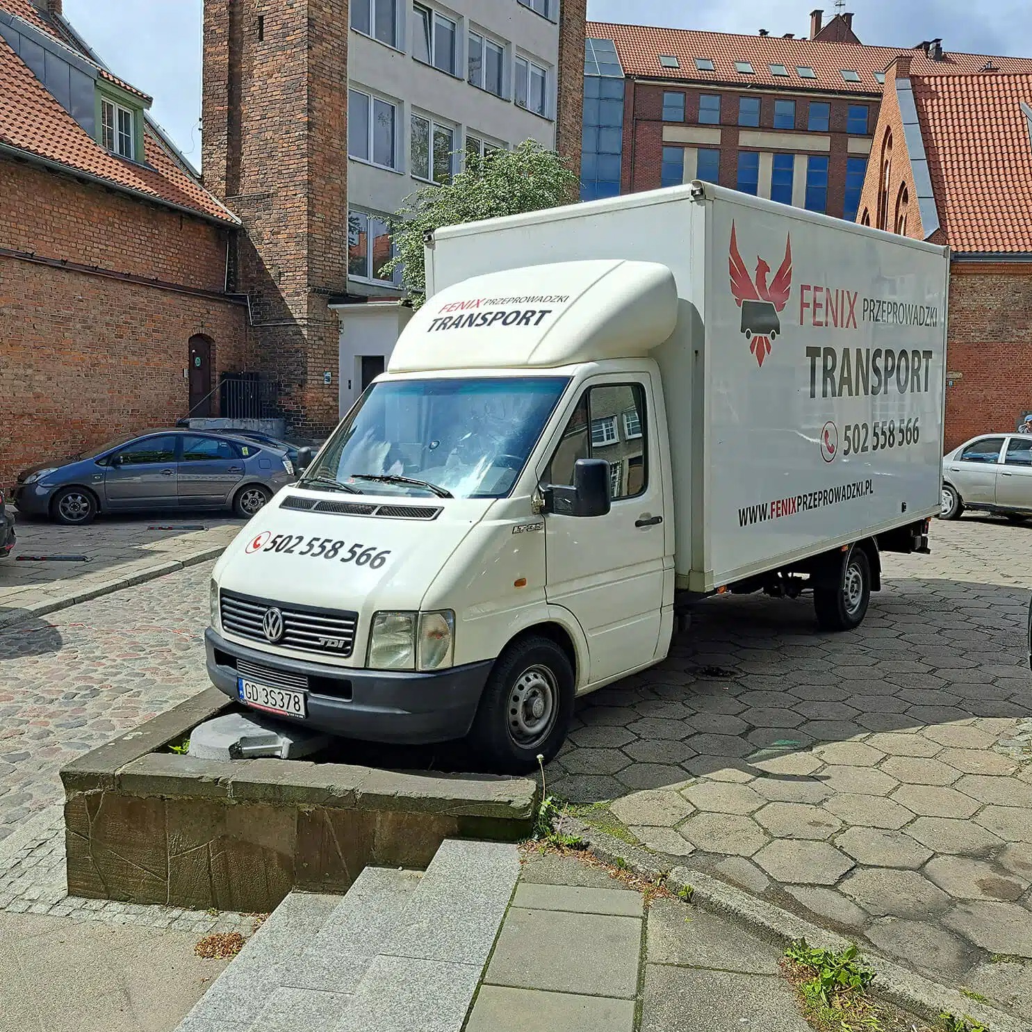 Biały samochód dostawczy z logo Fenix Przeprowadzki Transport na tle ceglanych budynków i szarej kostki brukowej. Widoczne numery kontaktowe na pojeździe.