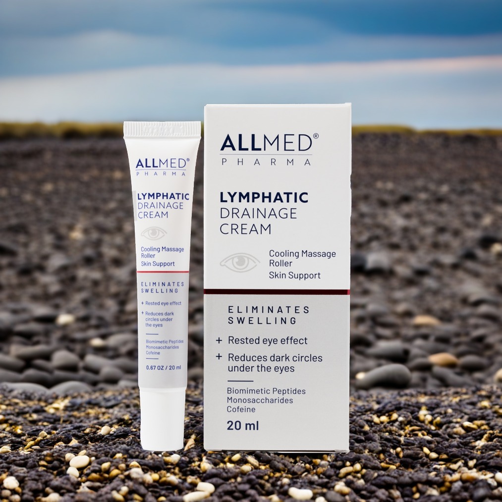 Krem Allmed Lymphatic Drainage w tubce i pudełku na tle kamienistej plaży pod błękitnym niebem. Produkt do pielęgnacji skóry, redukujący opuchliznę i cienie pod oczami.