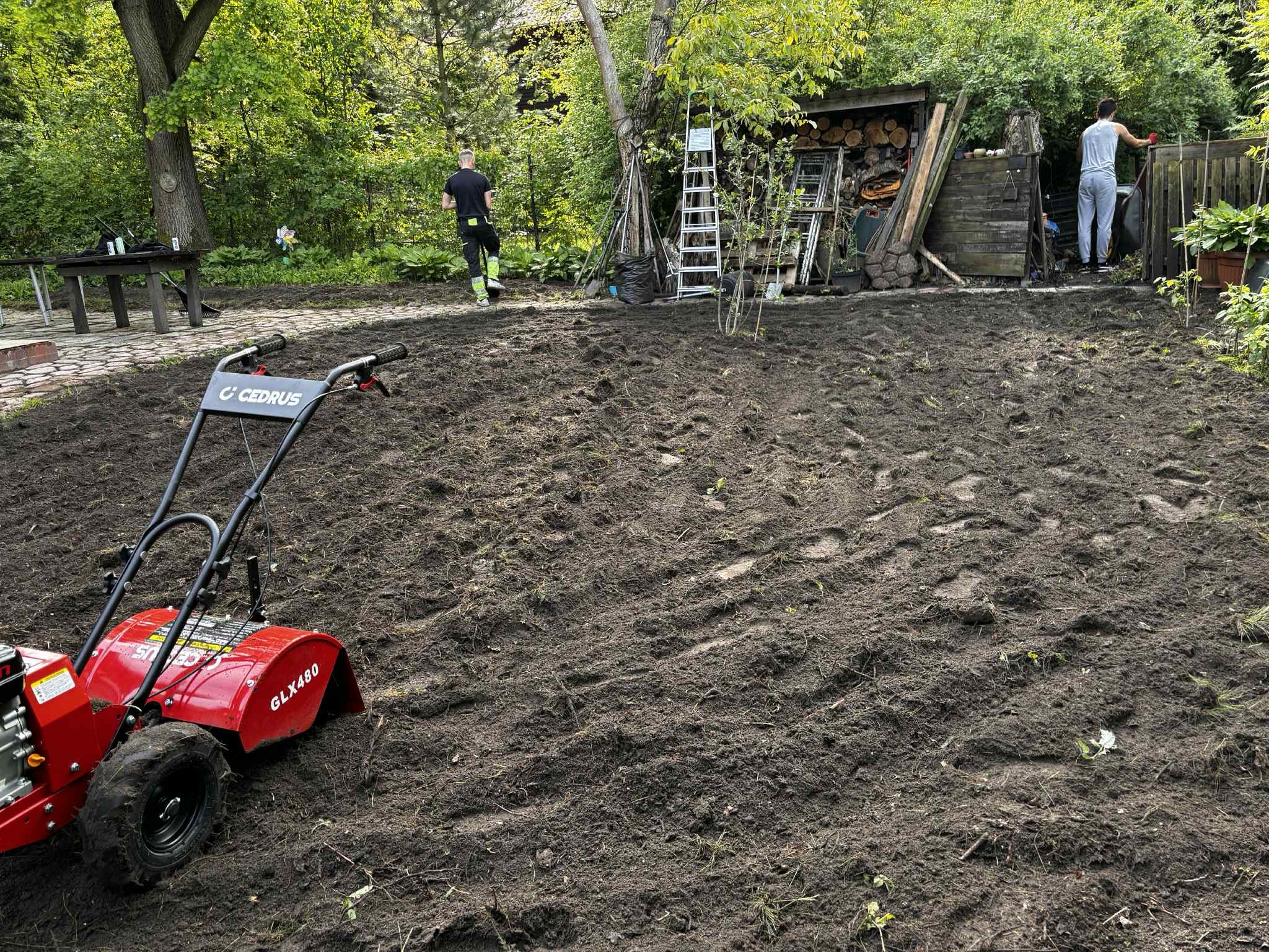 Przygotowany teren pod trawnik glebogryzarką Cedrus GLX480. Dwóch mężczyzn w tle. Ziemia przekopana, gotowa do siewu lub rozłożenia trawy z rolki. Ogród w tle.
