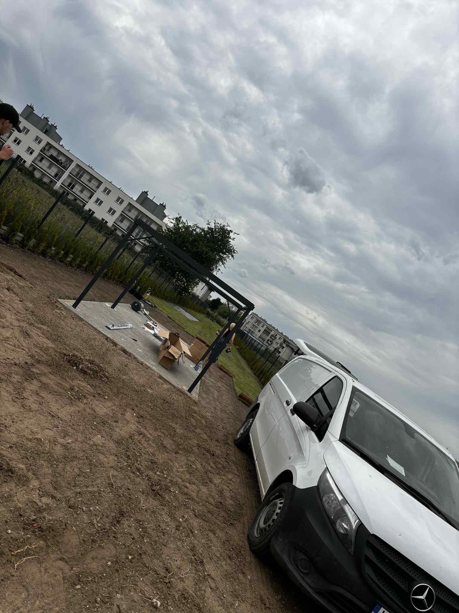 Montaż altany ogrodowej w Warszawie na przygotowanym podłożu, widoczny samochód dostawczy, pochmurne niebo.