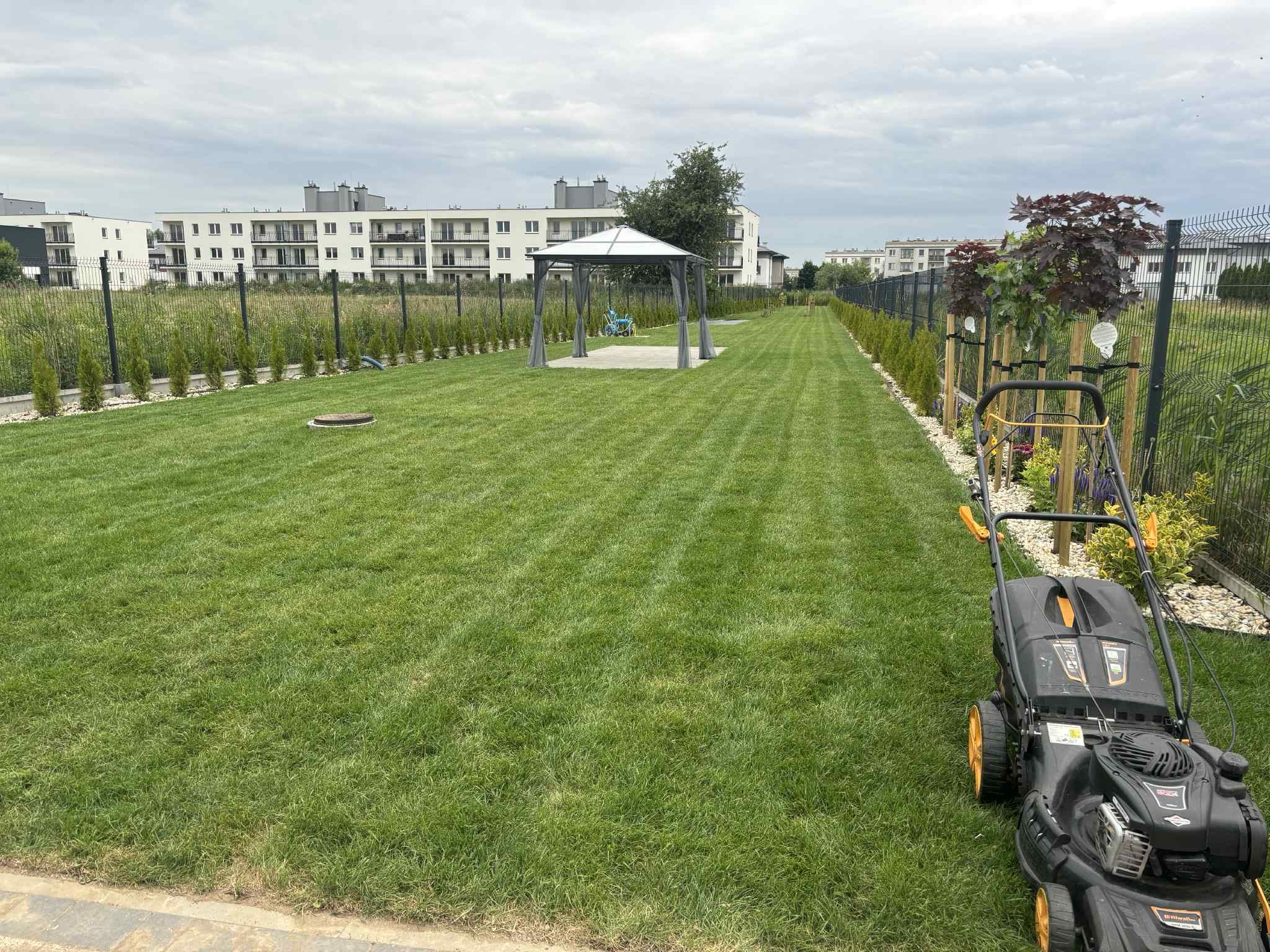 Równy trawnik po koszeniu z widocznymi pasami, na pierwszym planie kosiarka, w tle altana i budynki mieszkalne, obrzeża obsadzone tujami.