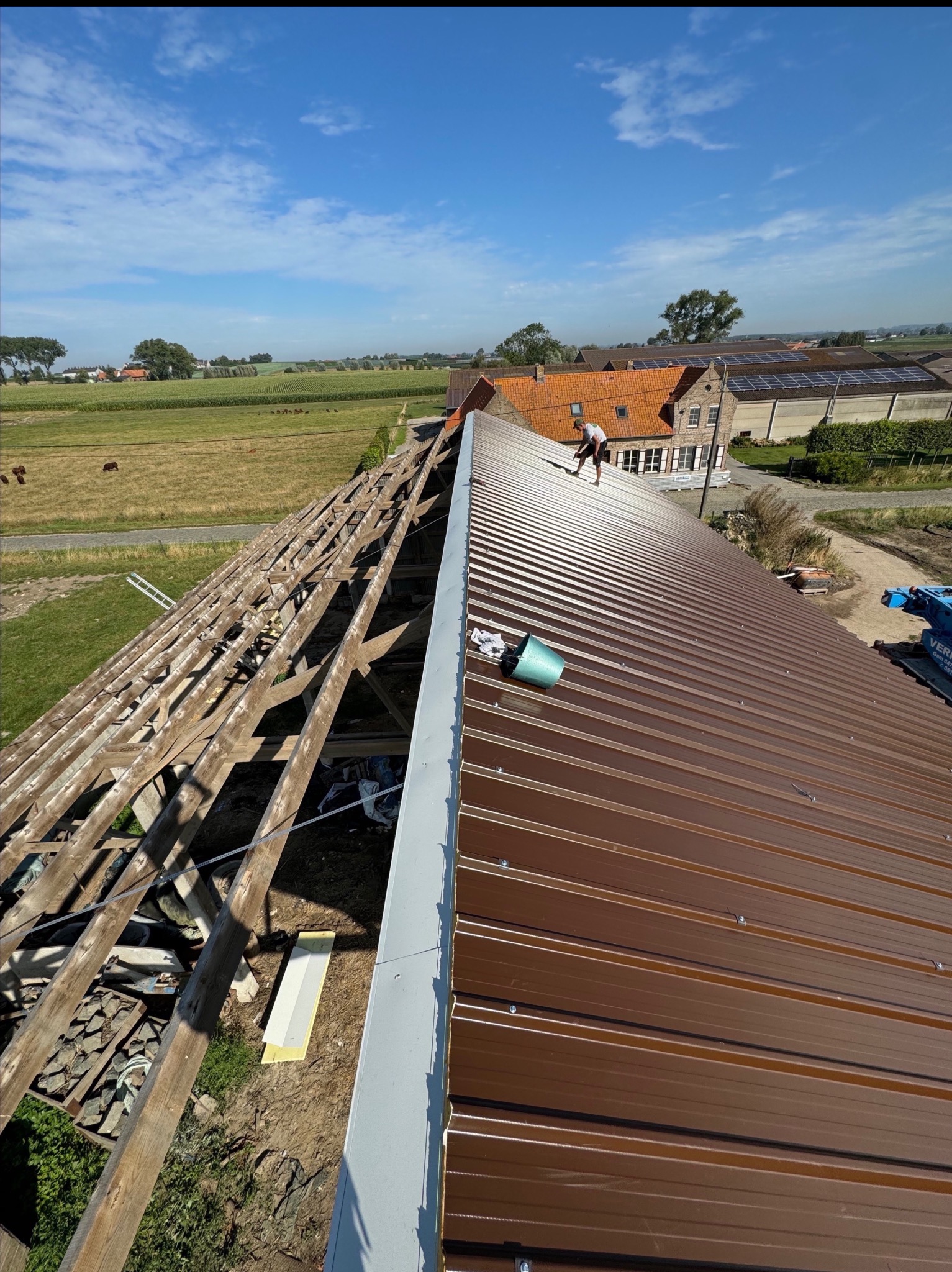 Montaż brązowego dachu z blachy trapezowej na drewnianej konstrukcji więźby, widok z góry na budynek gospodarczy z polami w tle, prace dekarskie w toku.