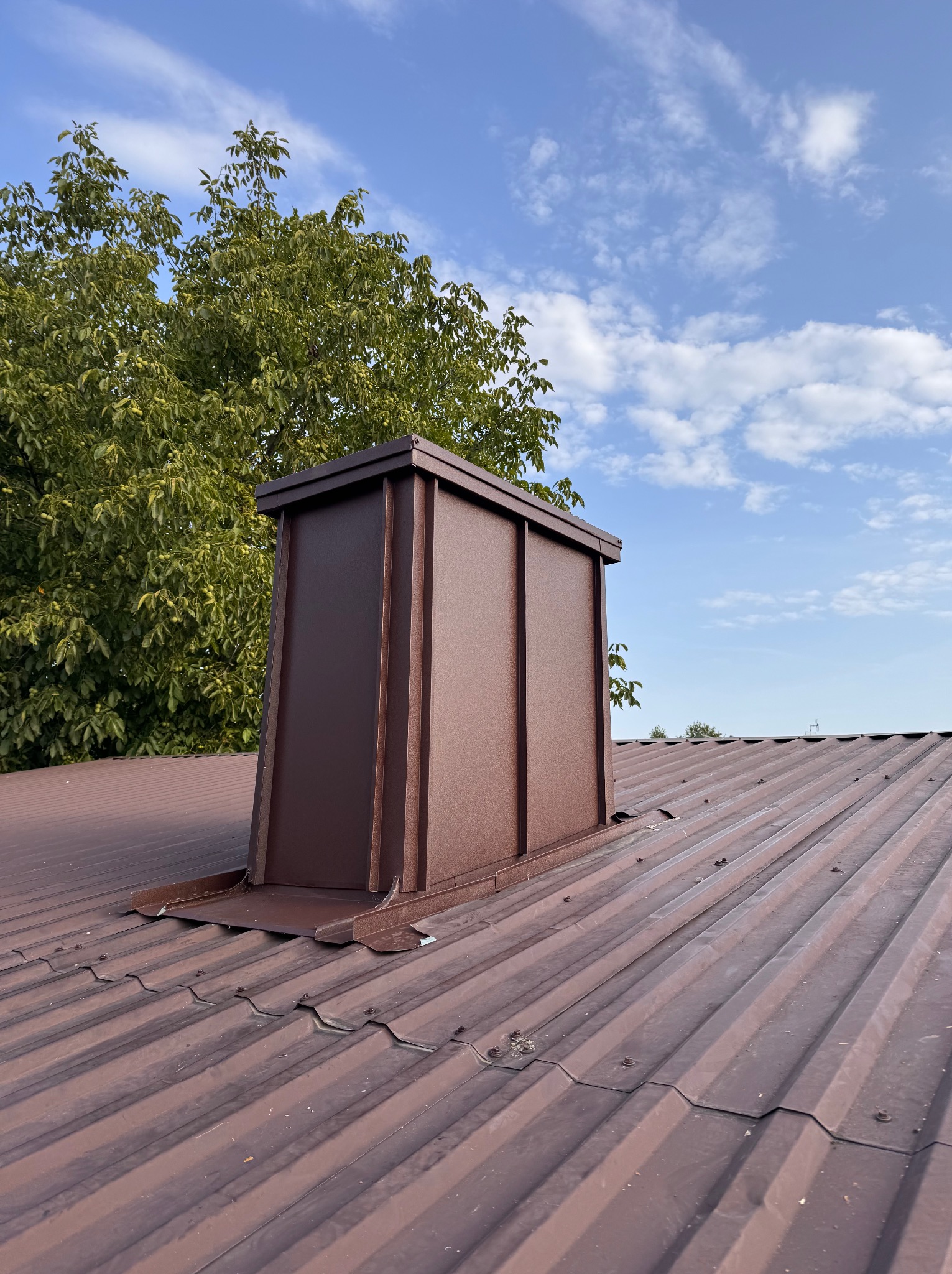 Brązowy komin wentylacyjny z blachy na dachu z blachy trapezowej, na tle błękitnego nieba i zielonych drzew. Widok z dołu, podkreślający detale obróbki blacharskiej.
