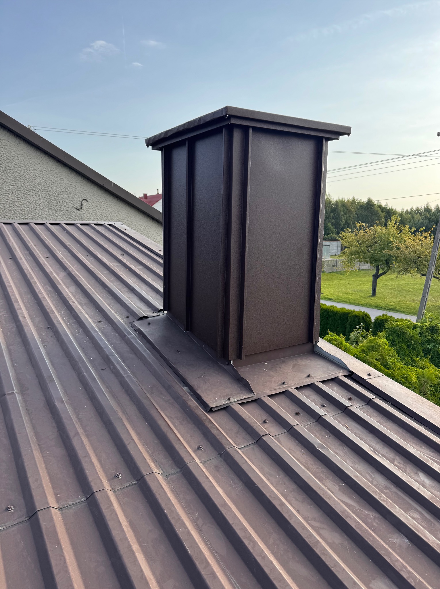 Nowy, brązowy komin wentylacyjny na dachu z blachy trapezowej. Komin z prostokątną obudową i daszkiem. Widok na zielone otoczenie i błękitne niebo.