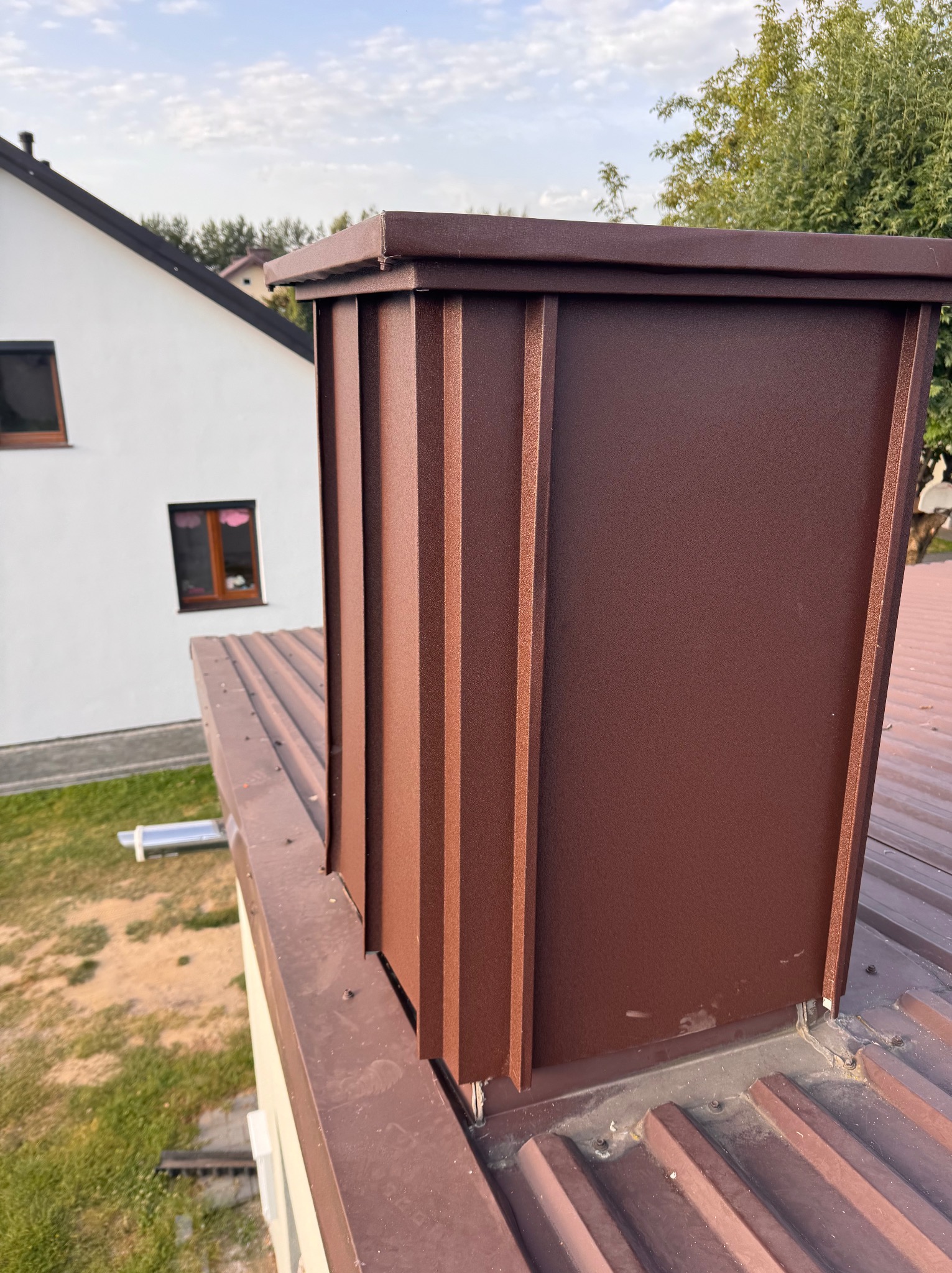 Brązowa obróbka blacharska komina na dachu z blachy trapezowej. W tle biały budynek z oknami i fragment zieleni. Widok z góry, oświetlenie dzienne.