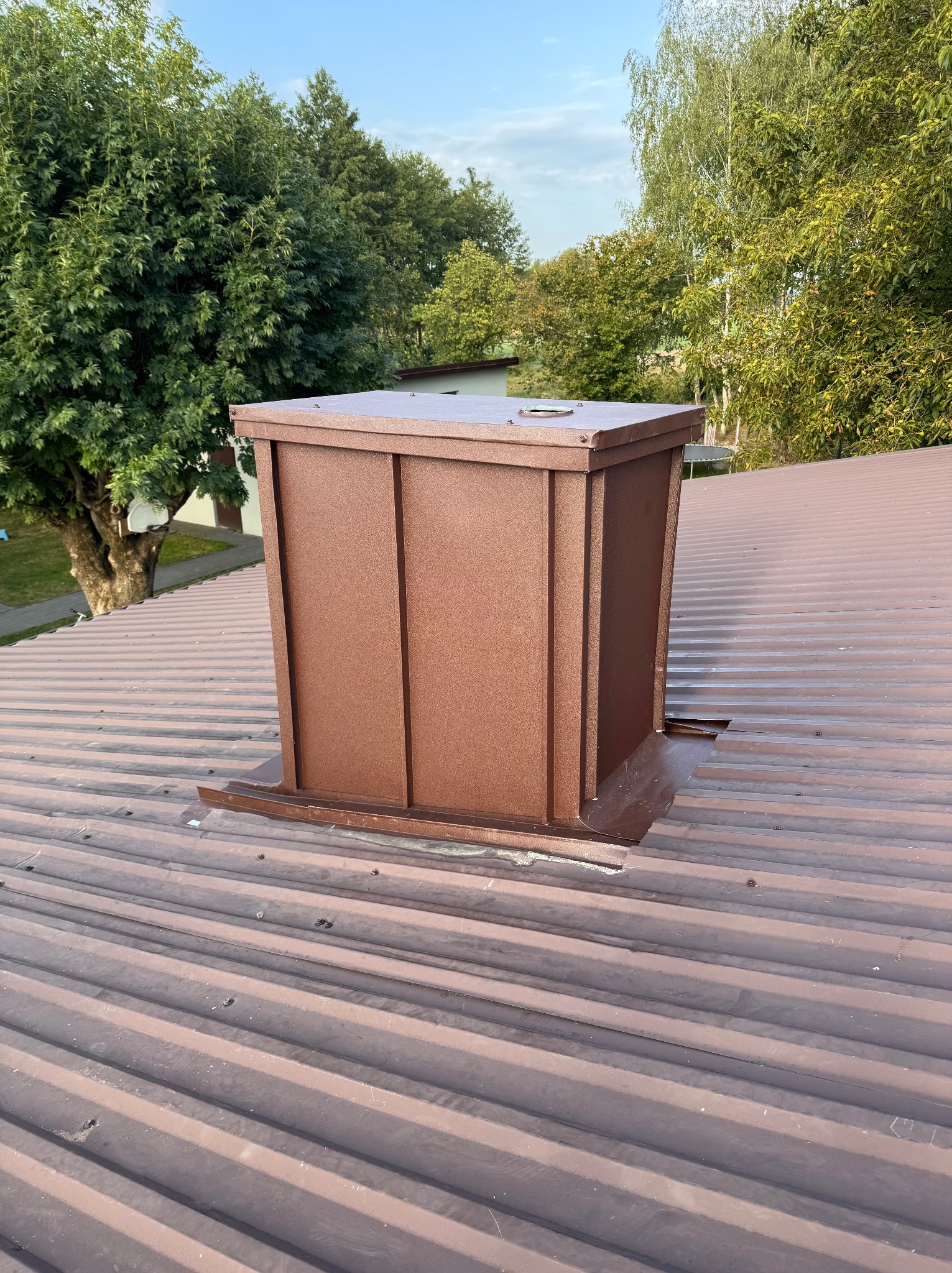 Brązowy komin z blachy na brązowym dachu z blachy trapezowej. W tle drzewa i błękitne niebo.