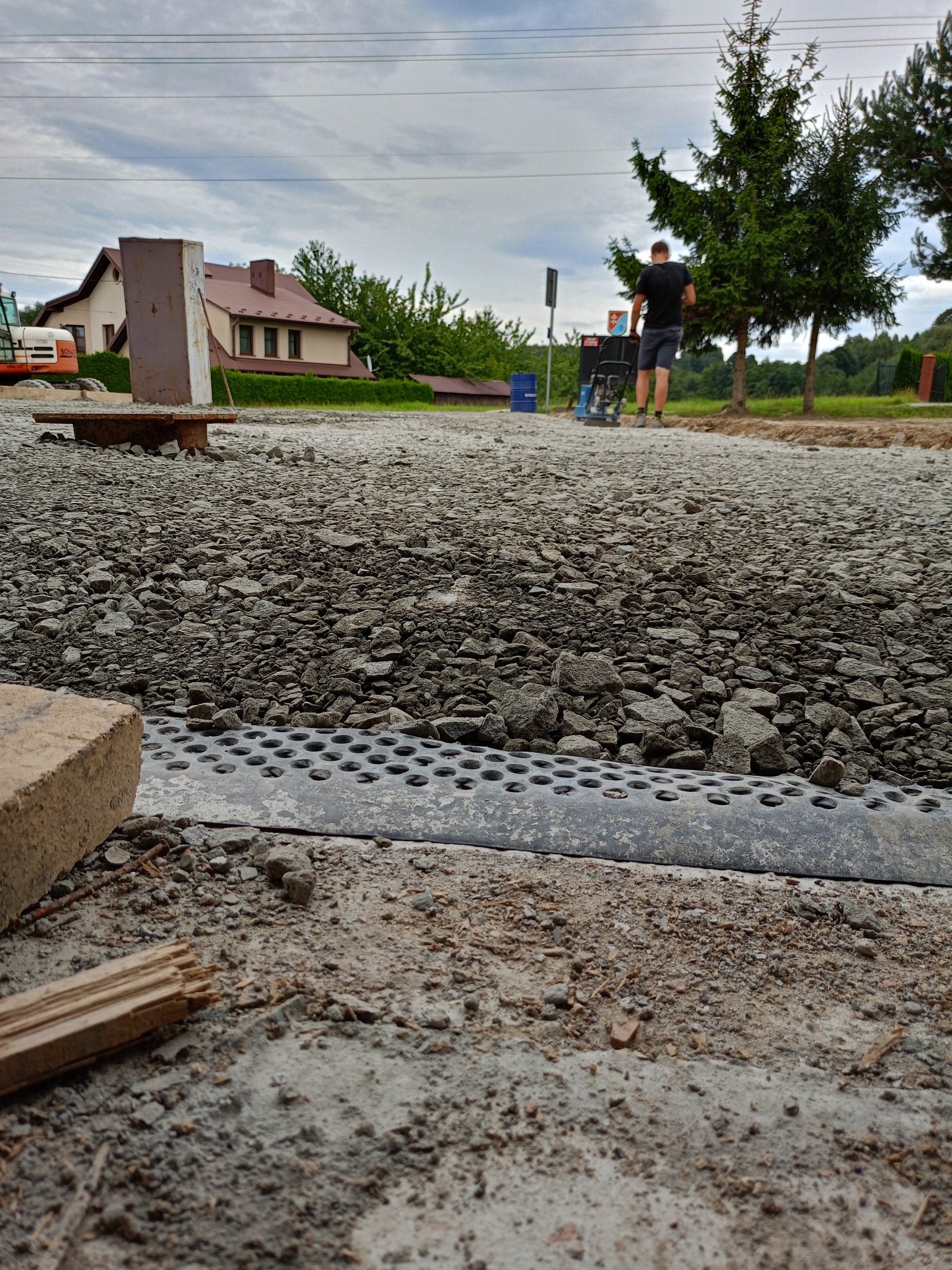 Utwardzanie terenu: ubijarka w akcji na żwirowej drodze z widocznym drenażem, dom w tle. Prace drogowe w toku, z bliska widoczny element drenażu.