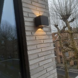 Damian Pucha - Kwadratowa, grafitowa lampa elewacyjna LED, zamontowana na ceglanej ścianie budynku. Ciepłe światło oświetla elewację. W tle widoczne drzewa i budynki.