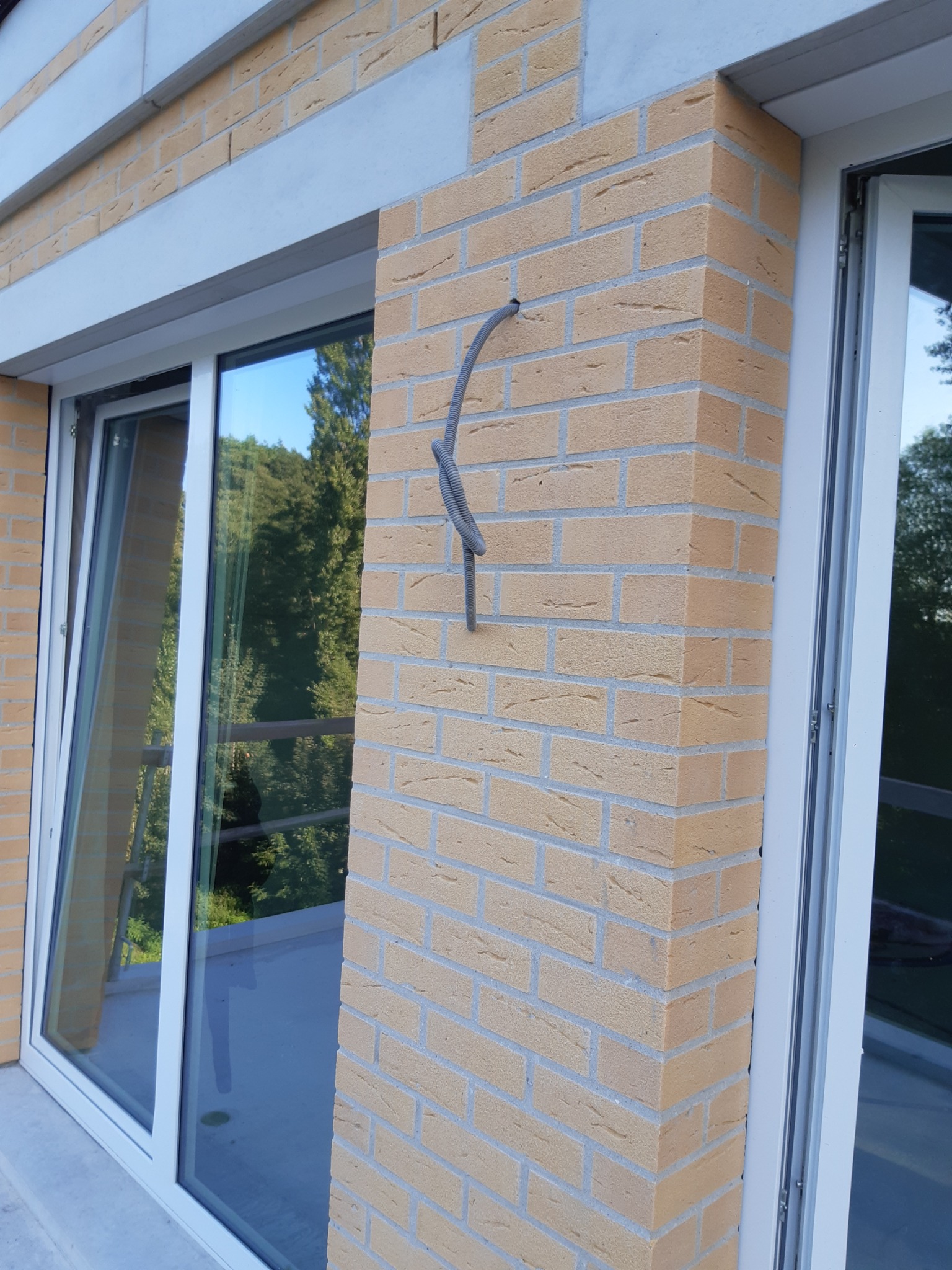 Brązowy mur z klinkieru przy oknie, z wystającym szarym przewodem elektrycznym. Widoczne drzewa w odbiciu szyby okiennej i biała rama okienna.