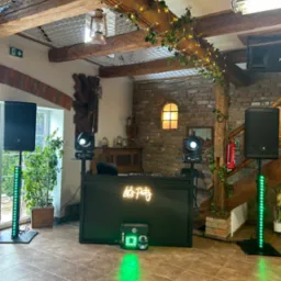 Dekoracja sali z konsoletą DJ-ską, oświetleniem punktowym i kolumnami głośnikowymi, neon 'Let's Party', rustykalne elementy wystroju.