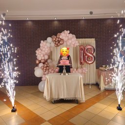 NAMAX EVENTS -DJ WODZIREJ FOTOBUDKA360 - Uroczystość 18-tych urodzin z tortem, balonami w odcieniach różu i złota oraz fontannami iskier po bokach. Dekoracja sali w ciepłej kolorystyce.