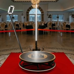 NAMAX EVENTS -DJ WODZIREJ FOTOBUDKA360 - Rotacyjna fotobudka 360 na czerwonym dywanie w eleganckiej sali balowej z ustawionymi stołami. Oświetlenie LED. Sprzęt gotowy do użycia.