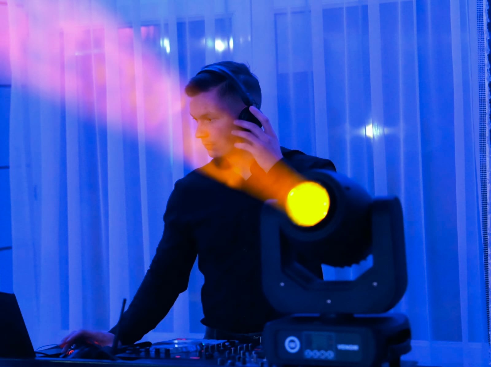 DJ w słuchawkach, oświetlony pomarańczowym światłem reflektora, obsługuje konsoletę w niebieskim oświetleniu. W tle białe zasłony.