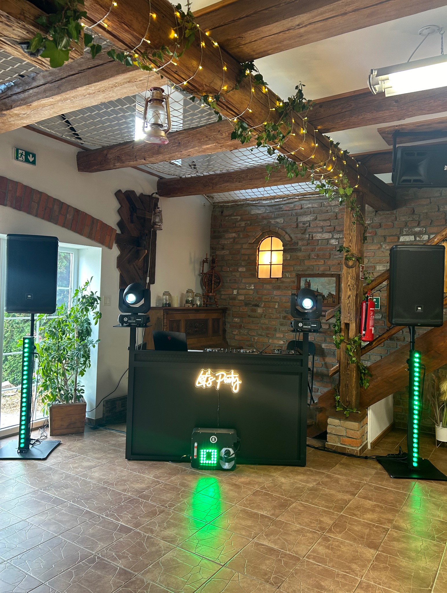 Dekoracja sali z konsoletą DJ-ską, oświetleniem punktowym i kolumnami głośnikowymi, neon 'Let's Party', rustykalne elementy wystroju.