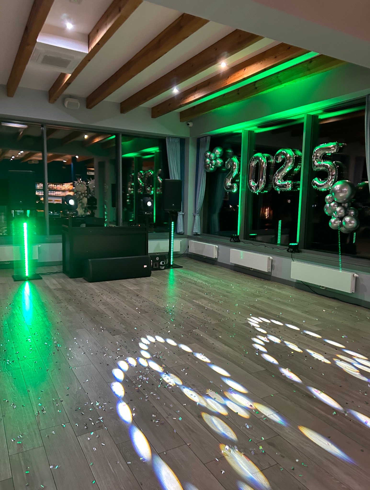 Sala z nagłośnieniem, oświetleniem i dekoracjami sylwestrowymi. Zielone światło podkreśla balony z napisem 2025, a konfetti leży na parkiecie.