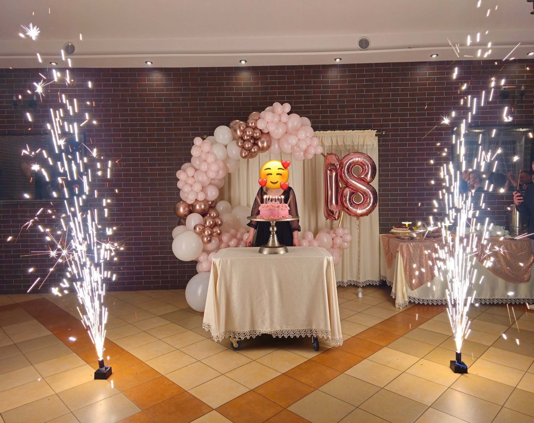 Uroczystość 18-tych urodzin z tortem, balonami w odcieniach różu i złota oraz fontannami iskier po bokach. Dekoracja sali w ciepłej kolorystyce.