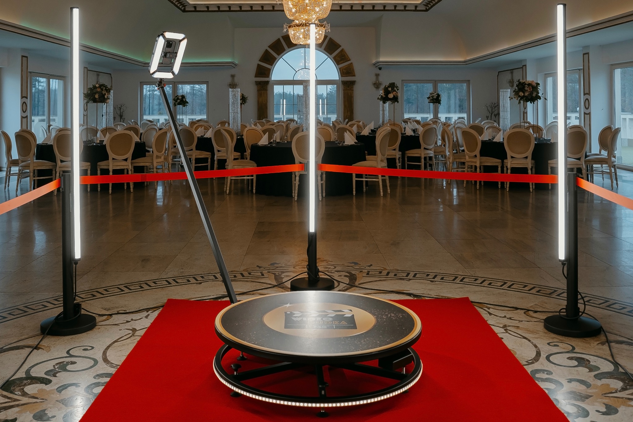 Rotacyjna fotobudka 360 na czerwonym dywanie w eleganckiej sali balowej z ustawionymi stołami. Oświetlenie LED. Sprzęt gotowy do użycia.