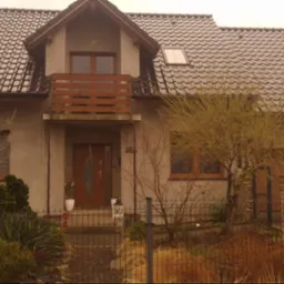 Dom jednorodzinny z szarą elewacją i ciemną dachówką. Widoczny balkon z drewnianą balustradą, okna z drewnianą ramą i zadbany ogród z metalowym ogrodzeniem.