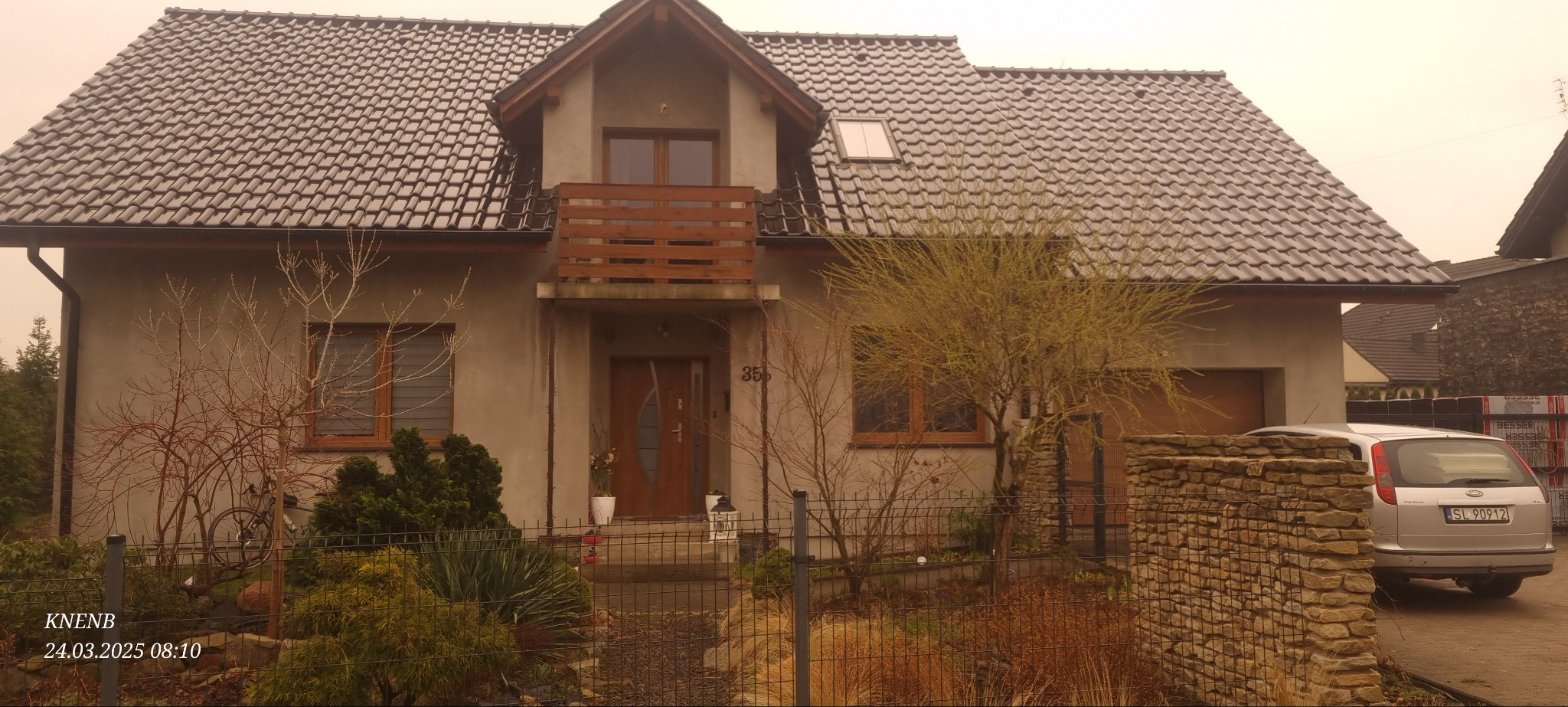 Dom jednorodzinny z szarą elewacją i ciemną dachówką. Widoczny balkon z drewnianą balustradą, okna z drewnianą ramą i zadbany ogród z metalowym ogrodzeniem.