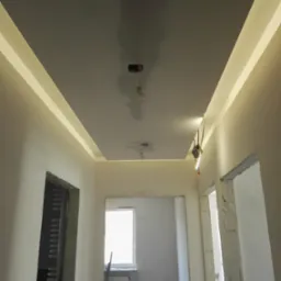 Wykończony sufit podwieszany z oświetleniem LED w korytarzu, widoczne puszki elektryczne, ściany przygotowane do malowania, surowy otwór drzwiowy.
